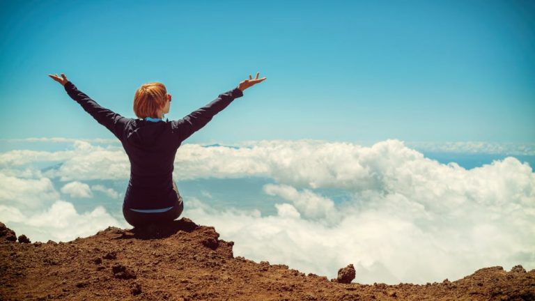 A woman enjoys a scenic view atop a cliff in Kula, Hawaii, surrounded by clouds and blue sky.